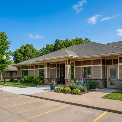 A single-story care center building with a gabled roof, brick and siding exterior, surrounded by landscaped gardens with colorful flowers, and a clear blue sky overhead.