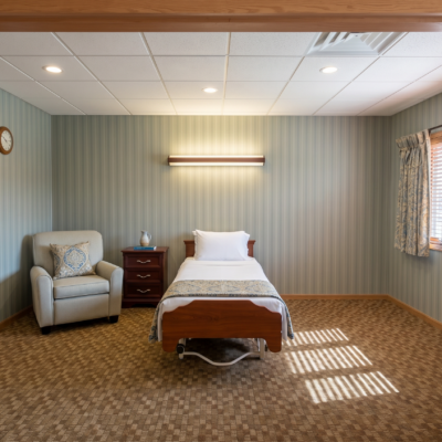 A bedroom with a single bed, a nightstand, and an armchair. The bed has white linens and a pillow. There is a window with patterned curtains on the right, letting in sunlight that creates a shadow on the carpet. A wall-mounted light fixture is above the bed, and a clock is on the left wall. The room has a beige patterned carpet, light green striped wallpaper, and a white ceiling with recessed lighting.