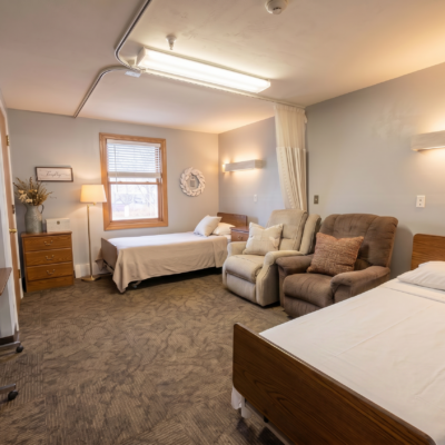 A spacious room with two beds, one with a white bedspread near a window with blinds, and the other with a white sheet and pillow. There are two armchairs, one beige and one darker, positioned between the beds. A wooden nightstand with a lamp and decorative items is next to the window. The room has soft lighting, light-colored walls, and a carpeted floor.