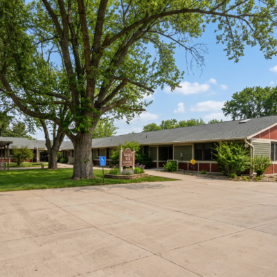 A single-story care center building with a landscaped front yard, large trees providing shade, and a parking lot in front on a sunny day with a blue sky and some clouds.
