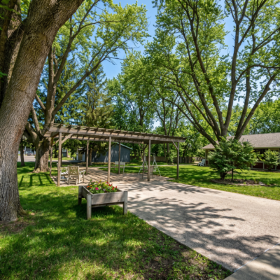 A shaded outdoor area with a wooden pergola, surrounded by lush green trees and grass. There is a small flower bed in a wooden planter and a bench nearby, with a house or building visible in the background under a clear blue sky.