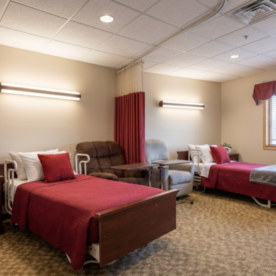A shared room in a care center with two beds, each covered with red blankets and pillows. There are two armchairs between the beds, a window with a red valance, and beige walls with horizontal light fixtures.