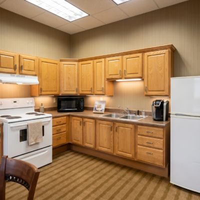A compact kitchen with wooden cabinets, a white stove, microwave, and refrigerator. There is a sink under the cabinets with a coffee maker on the counter nearby. The room has beige walls and a striped carpet.