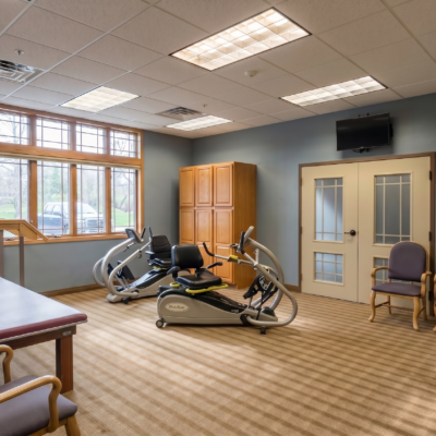 A room with large windows, exercise equipment including a recumbent bike and elliptical, wooden cabinet, wall-mounted TV, and chairs, suggesting a fitness or activity area in a care center.