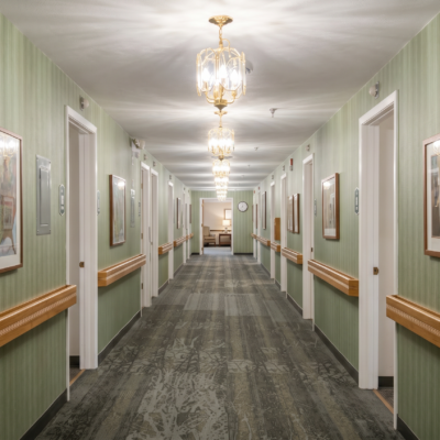 A long, well-lit hallway in a care center with green striped wallpaper, wooden handrails on both sides, framed pictures hanging on the walls, and elegant chandeliers hanging from the ceiling.