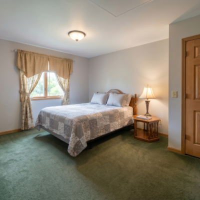 A bedroom with a bed covered in a patterned quilt, positioned against a white wall. There is a window with beige curtains and a nightstand with a lamp beside the bed. The room has green carpeted flooring and a wooden door.