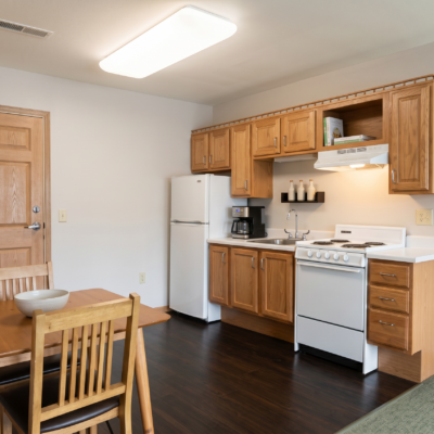 A small kitchen with wooden cabinets, a white refrigerator, a coffee maker, a stove, and a sink. There is a dining table with wooden chairs in the foreground.
