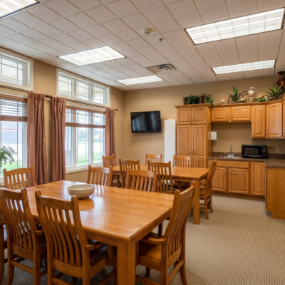 A dining area with a large wooden table surrounded by multiple matching chairs, next to a kitchenette with wooden cabinets, a microwave, and a small TV mounted on the wall. Large windows with curtains let in natural light.