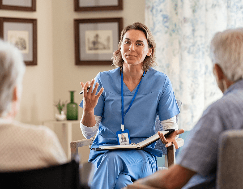 A female healthcare worker in blue scrubs with a name badge is sitting and talking to two elderly patients in a well-lit room with framed pictures on the wall. She is holding a notebook and a pen, gesturing with her hand as she speaks.