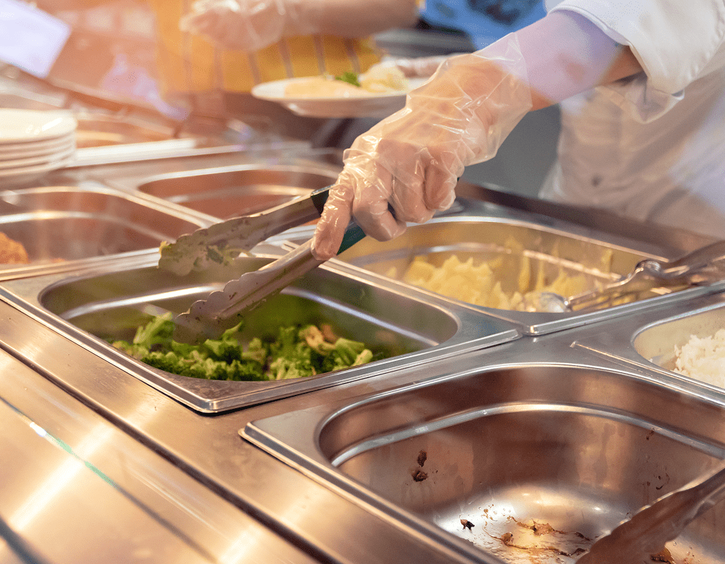 A person wearing a white coat, gloves, and using tongs is serving broccoli from a stainless steel cafeteria tray. Other trays with food, such as mashed potatoes and rice, are visible nearby.