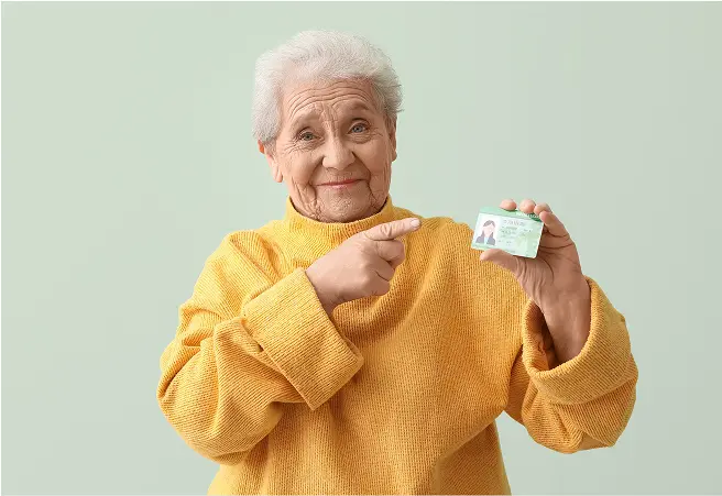 An elderly woman with gray hair, wearing a yellow sweater, smiling and pointing at her driver’s license which she holds in her right hand against a light green background.