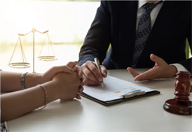 A person in a suit and tie is sitting at a table, signing a document with a gold pen, while another person holds their hands. In the background, there is a blurred image of a balance scale, symbolizing justice.