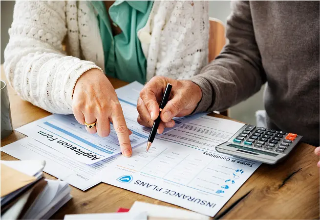 A person points to an insurance claim form while another holds a pen, with a calculator nearby; they are seated at a table with various papers and documents related to insurance and healthcare.