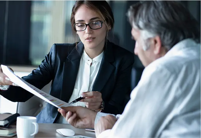 A young woman in business attire and glasses discusses documents with an older man in a white shirt at a table, in a professional setting.