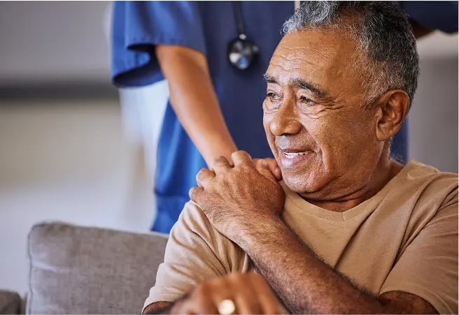A smiling elderly man sitting on a couch with a healthcare worker in blue scrubs standing behind him. The man is touching his shoulder and appears to be in a comfortable, caring environment.