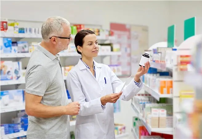 A female pharmacist showing a medication bottle to an older male customer inside a pharmacy with shelves stocked with various health and wellness products.